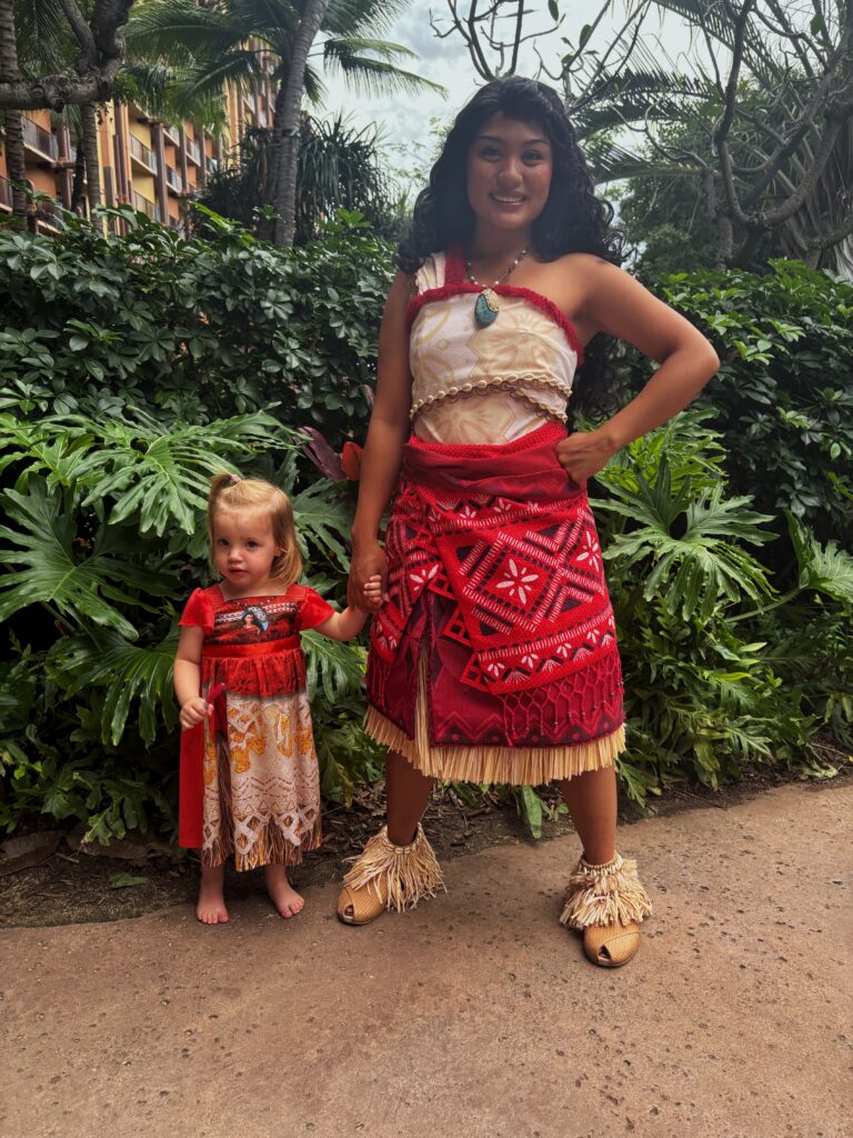 A young woman and a little girl stand outdoors, both dressed in matching red, tan, and white Polynesian-inspired costumes, holding hands and smiling amidst tropical greenery.
