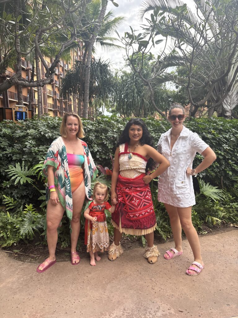 Three women and a young girl pose outdoors by lush greenery. One woman and the child wear tropical swimsuits, another wears a white cover-up, and a woman in the center wears a traditional red Polynesian outfit.