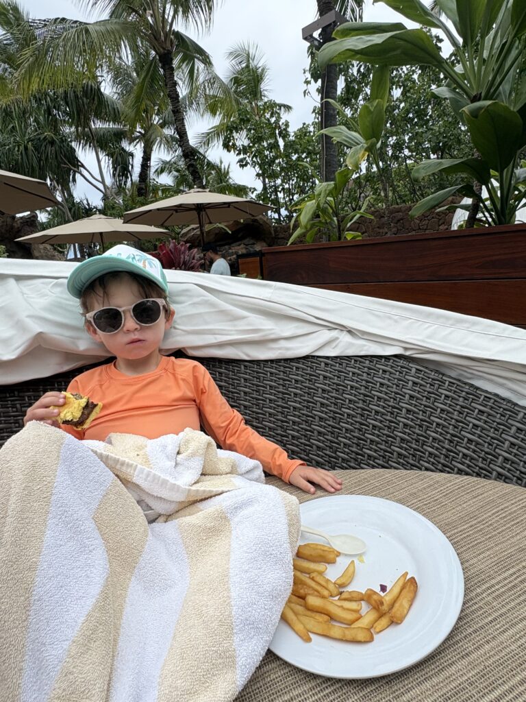 A young child in sunglasses and an orange shirt lounges on a wicker chair with a towel, eating a burger and fries. Palm trees and lush plants surround the outdoor setting.