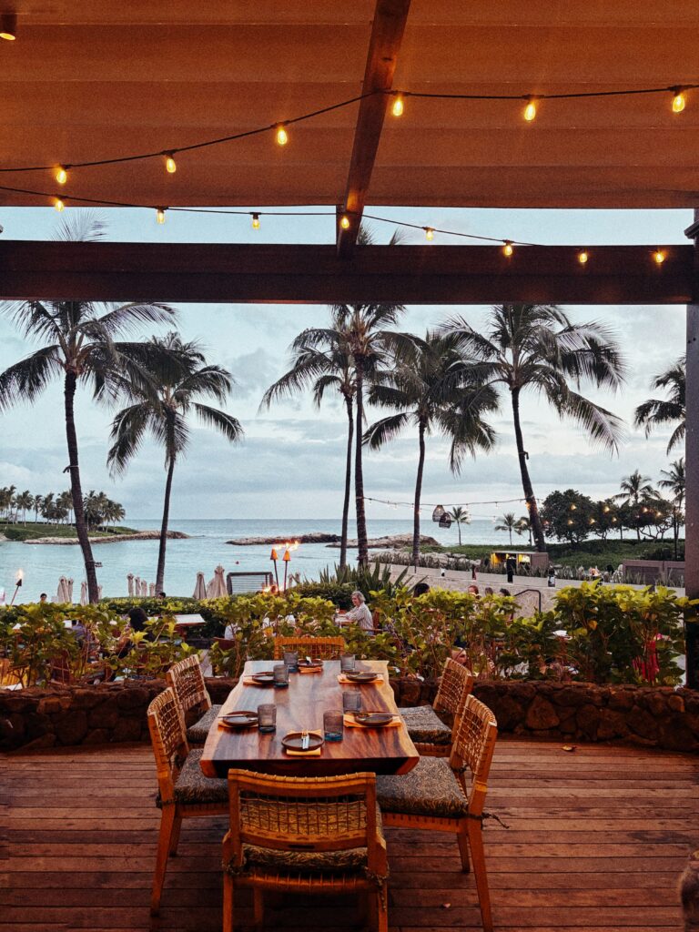 A wooden outdoor dining table set for four overlooks a beach with palm trees at sunset, string lights hanging above and the ocean visible in the background.