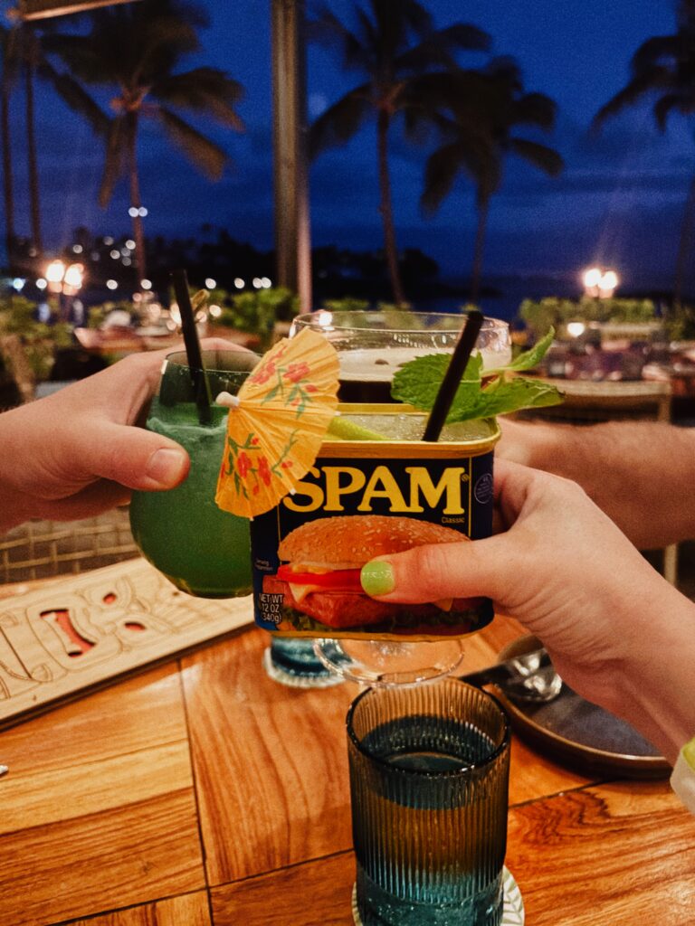 Two people clink colorful cocktails at an outdoor table at night. One drink is in a blue glass, the other is served in a SPAM can with an umbrella and mint. Palm trees and lights are visible in the background.