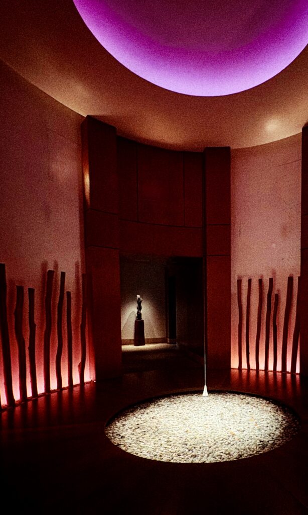 A dimly lit circular room with wooden columns, a central light illuminating a pebble-filled circle on the floor, and purple ceiling lighting; a statue is visible in an alcove at the back.