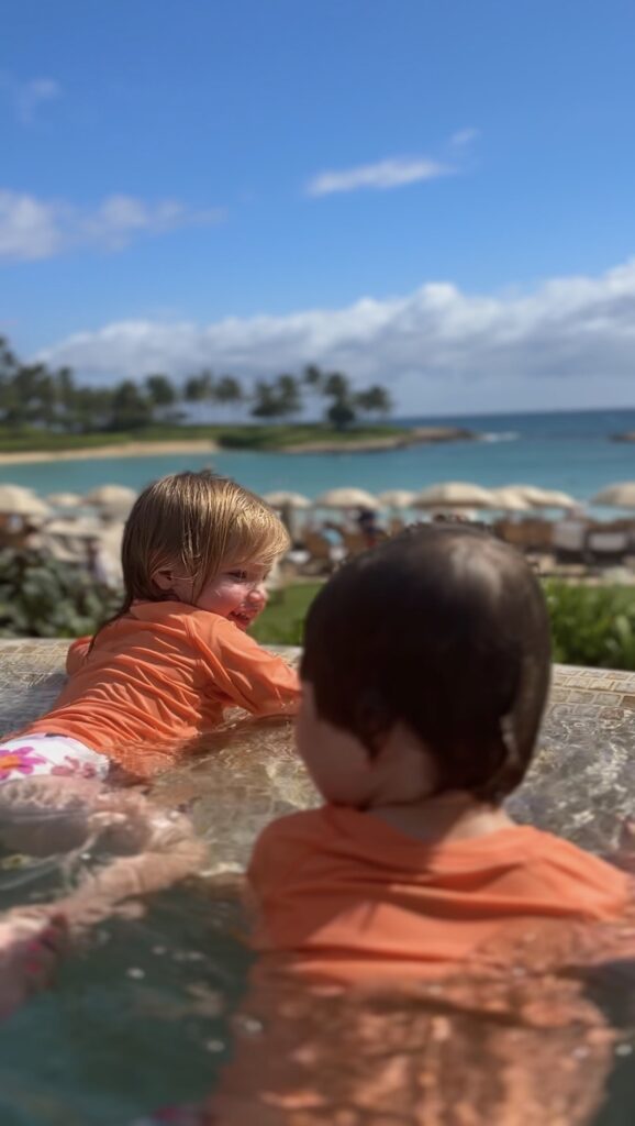 Two young children in orange swim shirts play in a shallow pool, smiling and looking toward the ocean. Beach chairs, umbrellas, palm trees, and a blue sky with clouds are visible in the background.