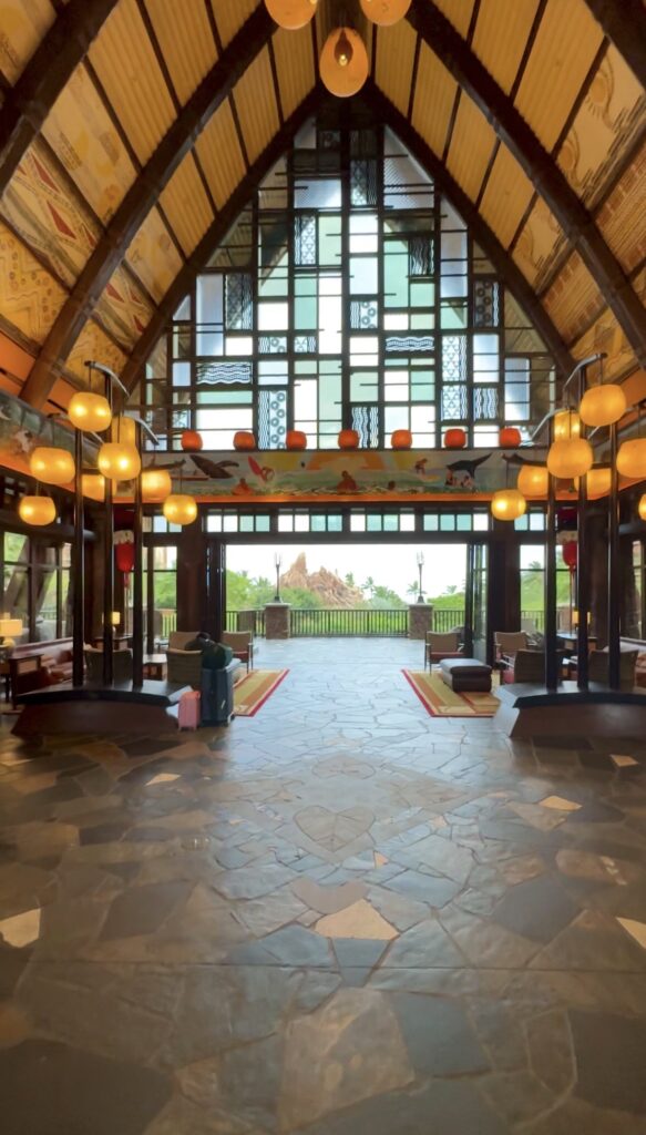 A spacious hotel lobby with high, vaulted ceilings, large windows, warm lighting, decorative patterns, and luggage near the entrance; outdoor scenery is visible through the glass.