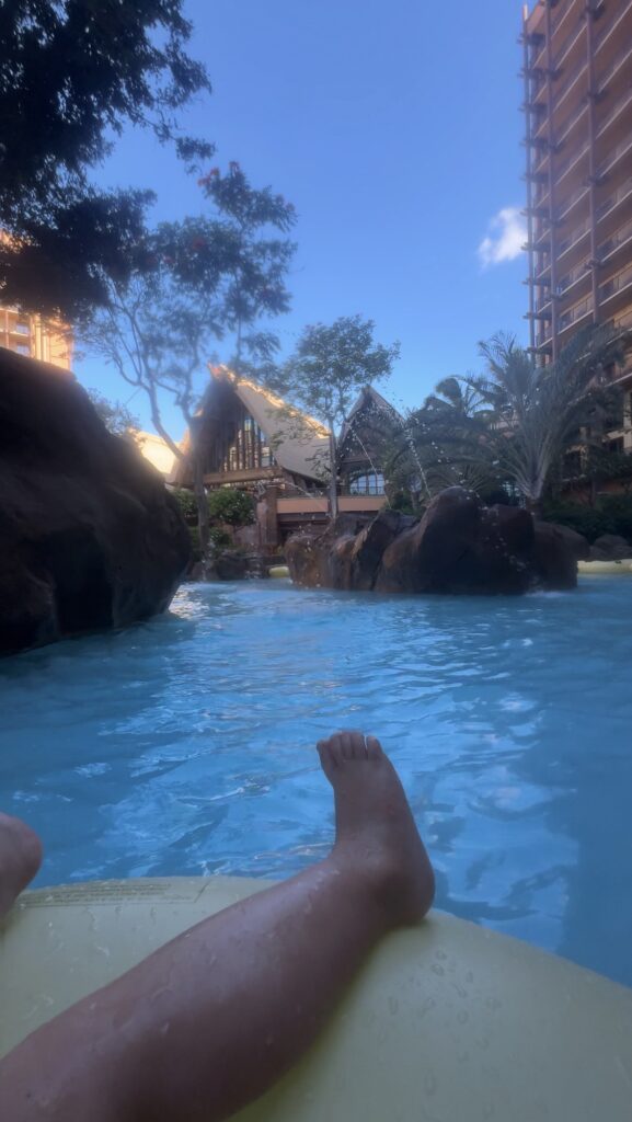 Person relaxing on a pool float, feet visible in the foreground, surrounded by rocks, trees, palm trees, and tall resort buildings under a blue sky. A tropical, tranquil vacation setting.