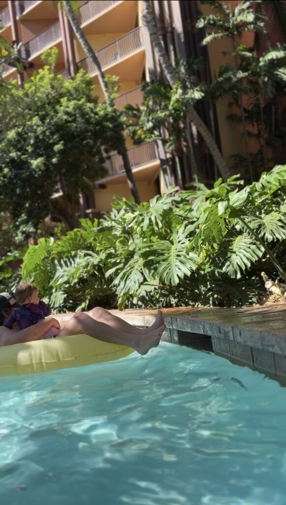 A person relaxes on a yellow float in a pool, leaning back with legs stretched out. Lush green plants and trees surround the pool, and a multi-story building is visible in the background.