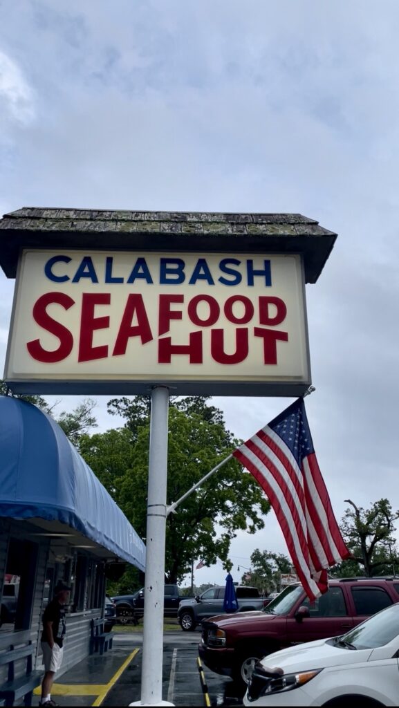 A sign reading Calabash Seafood Hut stands outside, with an American flag beside it. Part of a blue awning and building, along with trees and parked cars, are visible under a cloudy sky.