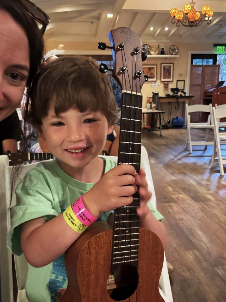 A smiling young child wearing a green shirt and event wristbands holds a small ukulele indoors. An adult is partially visible next to the child. The setting appears warm and welcoming with wooden floors and chairs.