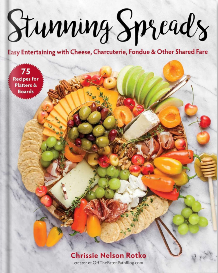 A cookbook titled Stunning Spreads with a cover featuring an elaborate platter of cheeses, grapes, apples, honey, nuts, crackers, and various colorful fruits and vegetables on a marble background—perfect inspiration for your own stunning spreads.