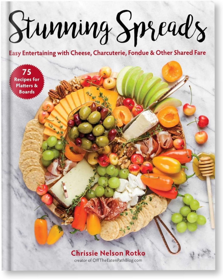 The cover of the book Stunning Spreads features an elaborate charcuterie board with cheeses, grapes, crackers, nuts, and honey, arranged on a marble surface. The book promises 75 recipes for platters and boards.