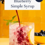 A glass of ice, lemon, and blueberries is being filled with blueberry syrup. A sprig of mint garnishes the drink. The background is yellow, with fresh blueberries and a lemon bowl visible. Text reads Blueberry Simple Syrup—So Easy!.