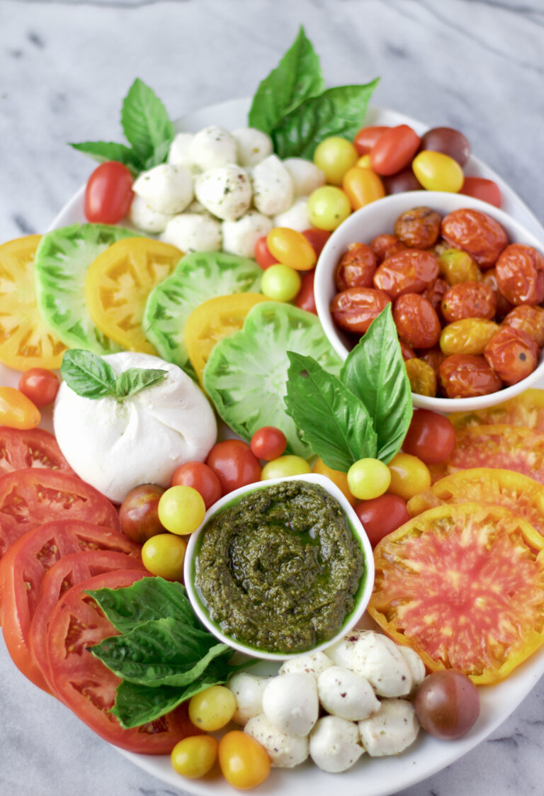 A colorful platter featuring heirloom tomato slices, cherry tomatoes, mozzarella balls, fresh basil, a bowl of roasted tomatoes, and a bowl of green pesto on a white marble background.