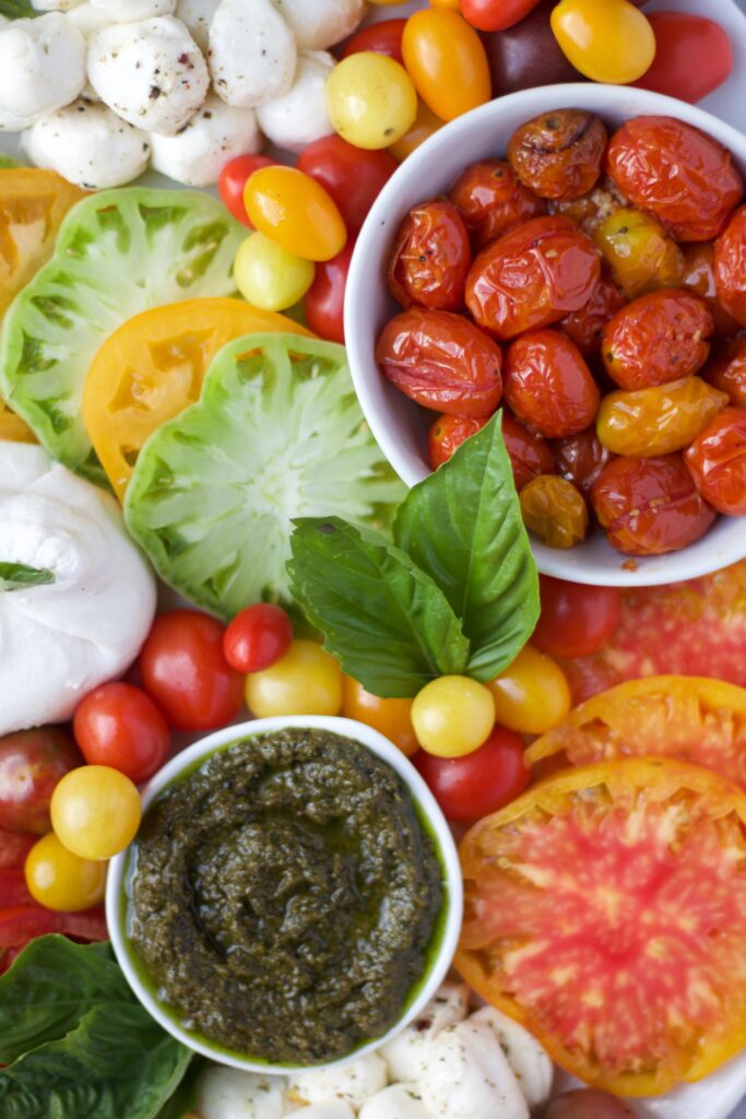A colorful assortment of heirloom tomato slices, cherry tomatoes, mozzarella balls, roasted tomatoes in a bowl, basil leaves, whole garlic, and a bowl of green pesto.