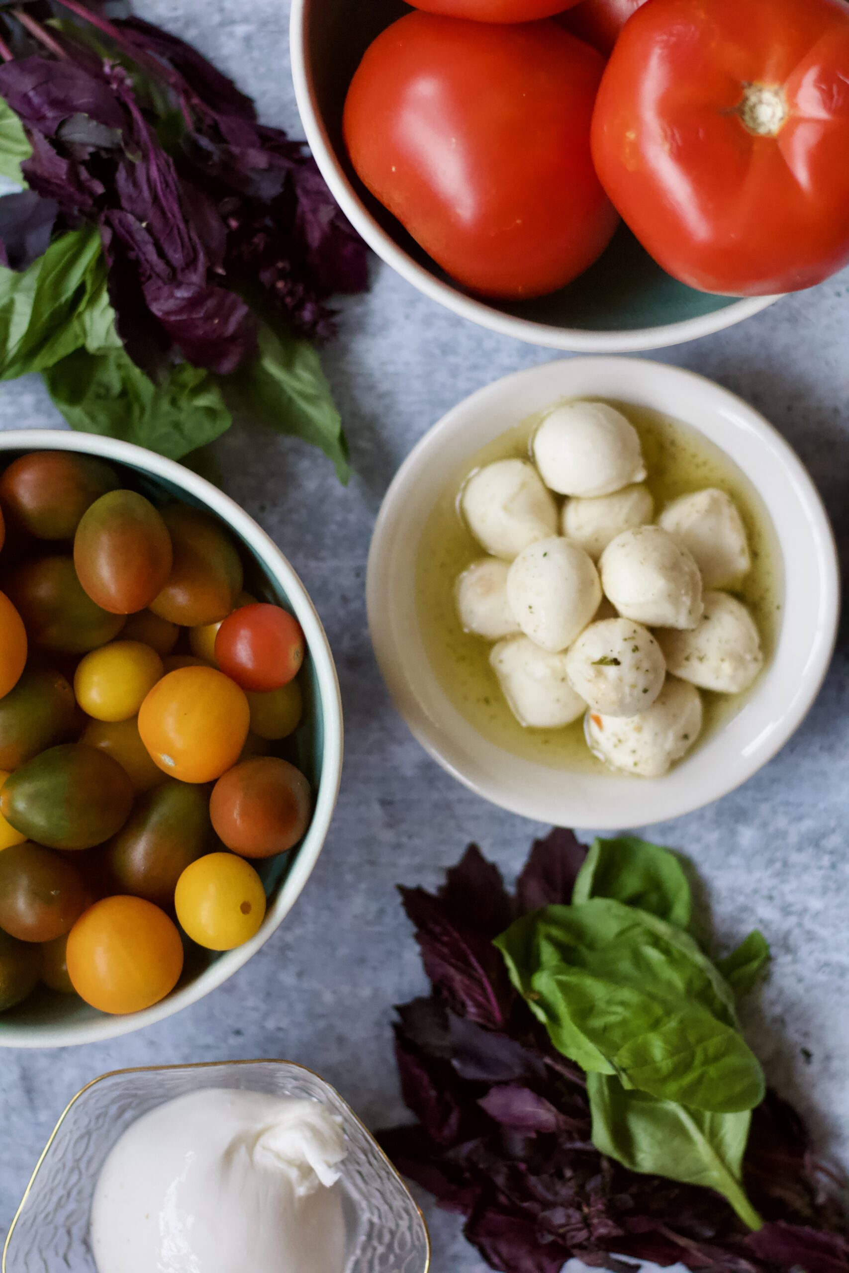 Bowls of red tomatoes, cherry tomatoes, mozzarella balls in oil, and fresh basil leaves arranged on a light blue surface.