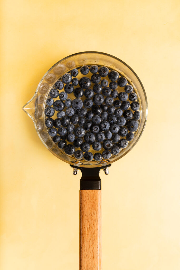 A glass saucepan filled with fresh blueberries sits on a pale yellow background, viewed from above. The saucepan has a light wooden handle.