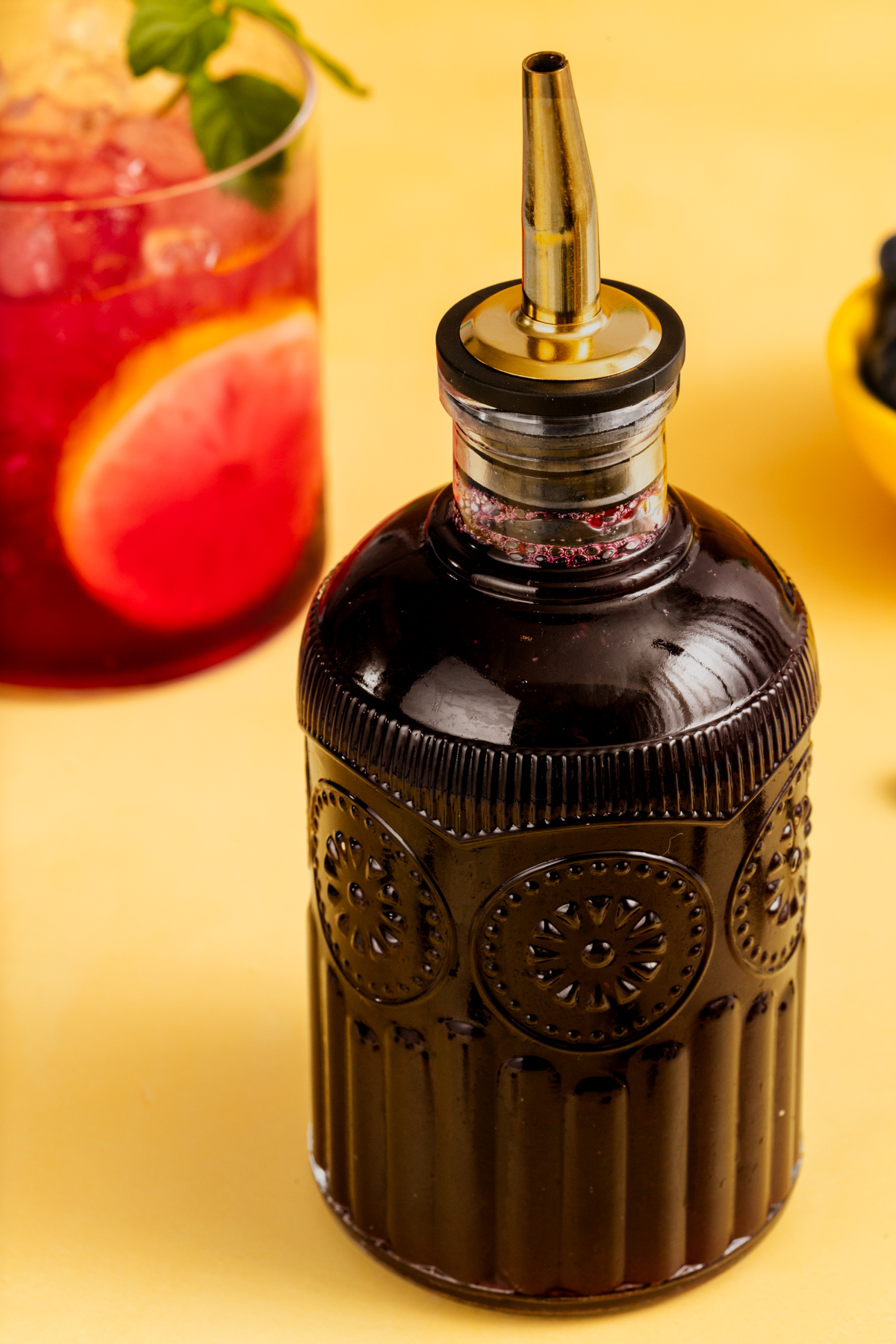A decorative glass bottle with dark liquid and a metal spout sits on a yellow surface, next to a red drink garnished with lemon and a small yellow bowl of blueberries in the background.