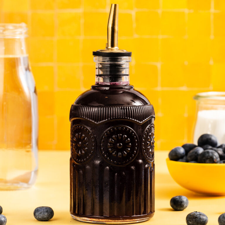 A decorative glass bottle with a metal spout, filled with dark blueberry syrup, sits on a yellow surface near a bowl of blueberries and a glass of water, against a yellow tiled background.
