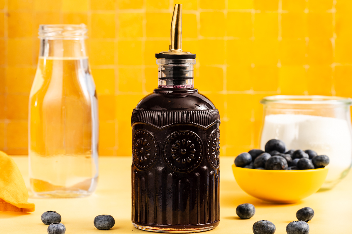A glass bottle of dark blueberry syrup with a metal pour spout stands in front of a yellow tiled wall, surrounded by a glass of water, a small yellow bowl of blueberries, and a jar of sugar.