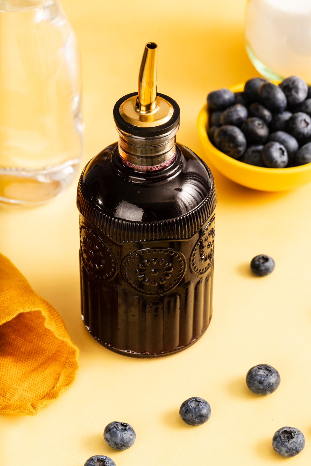 A glass bottle with a pour spout holds dark liquid, surrounded by scattered blueberries, a yellow cloth, a yellow bowl of blueberries, and a glass of water, all on a yellow surface.