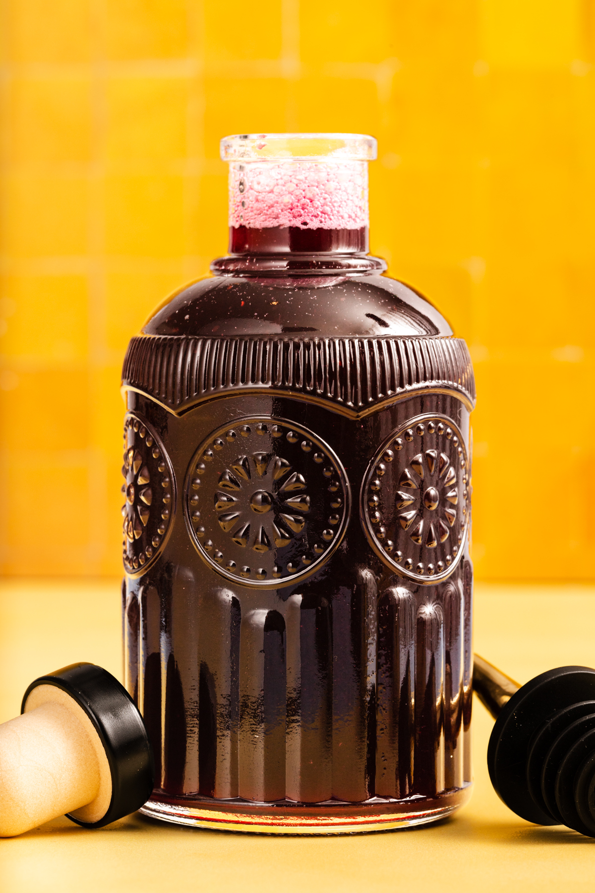 A decorative glass bottle filled with dark liquid and foam on top is flanked by two honey dippers, set against a bright yellow tiled background.