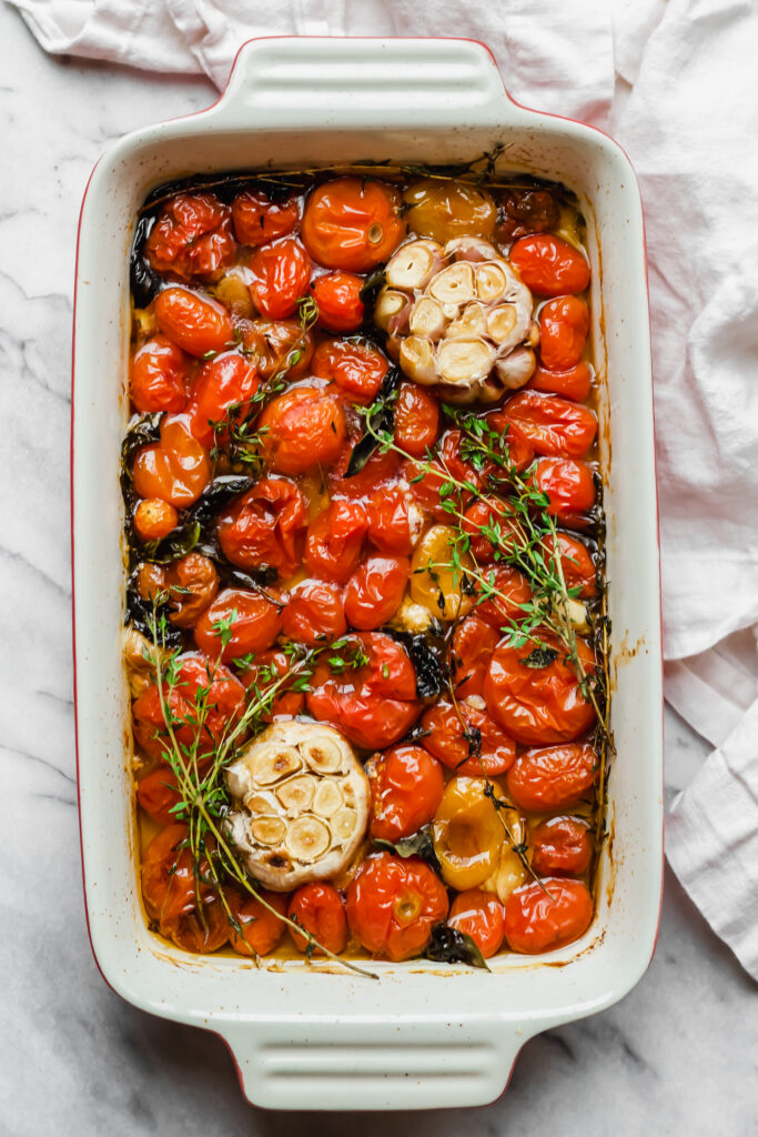 A white baking dish filled with roasted cherry tomatoes, two halved heads of garlic, sprigs of fresh thyme, and a light golden roasting liquid, set on a marble surface with a white cloth nearby.
