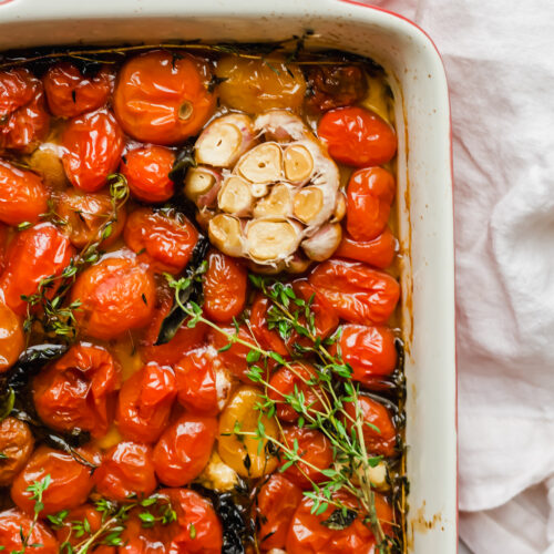 A roasting pan filled with baked cherry tomatoes, halved garlic bulbs, fresh thyme sprigs, and herbs on a white cloth background.