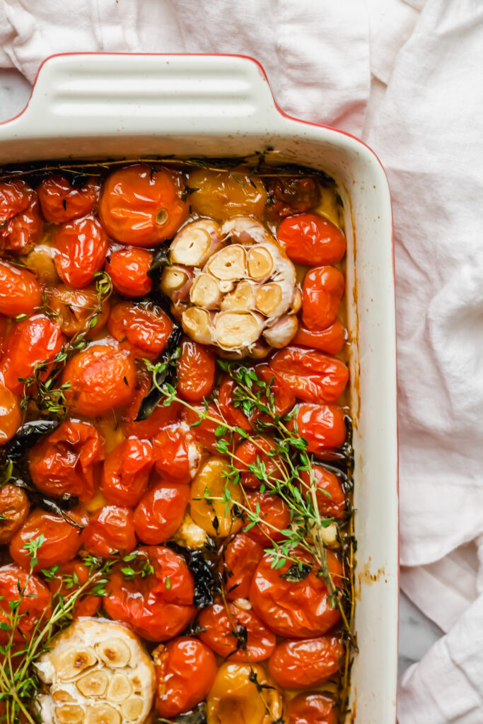 A roasting pan filled with baked cherry tomatoes, halved garlic bulbs, fresh thyme sprigs, and herbs on a white cloth background.
