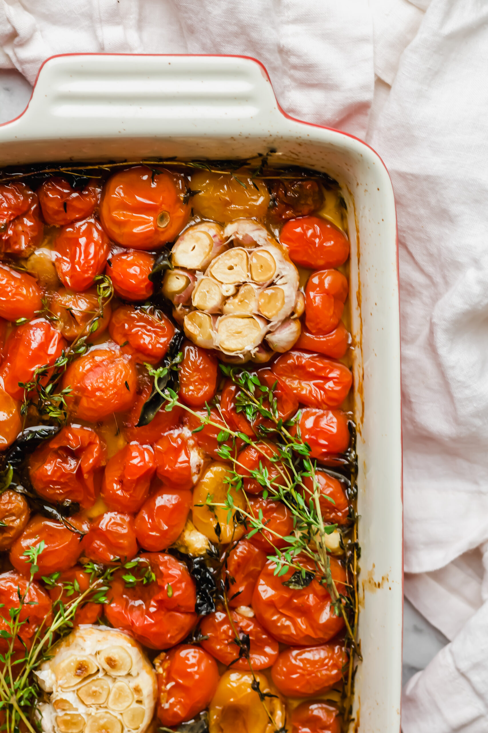 A roasting pan filled with baked cherry tomatoes, halved garlic bulbs, fresh thyme sprigs, and herbs on a white cloth background.