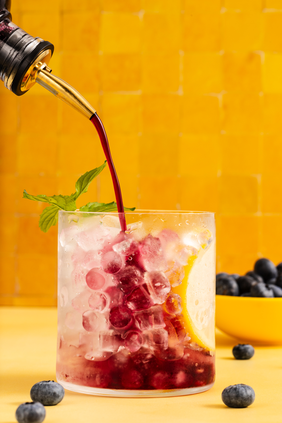 A glass of ice with lemon and mint is being filled with red liquid. Blueberries are in the drink and scattered on the yellow surface. The background is a yellow tiled wall.