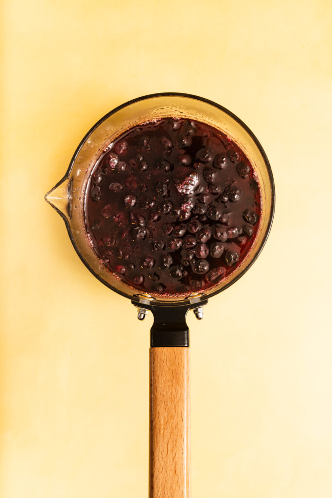 A glass saucepan with a wooden handle filled with simmering blueberry sauce, viewed from above, set against a yellow background.