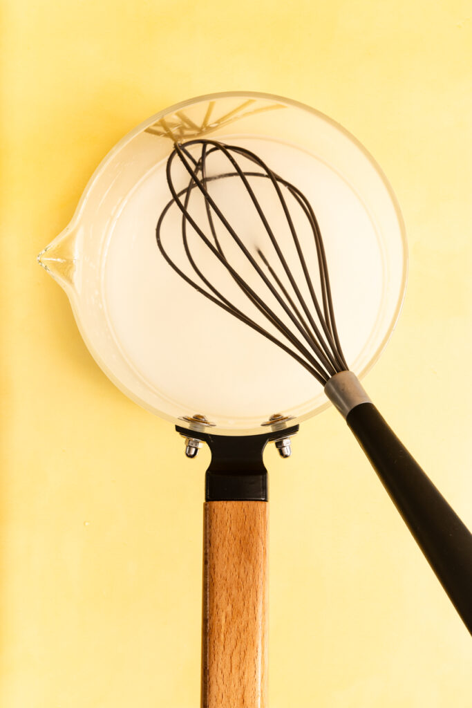 A glass saucepan with a wooden handle holds a white liquid being whisked with a black metal whisk, set against a bright yellow background.