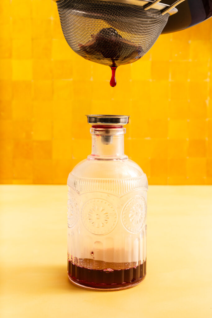 A dark liquid drips from a strainer into a clear, vintage-style glass bottle against a bright yellow background.