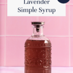 A glass bottle filled with lavender simple syrup sits on a pink counter with a pink tile backdrop. The label reads Lavender Simple Syrup and includes a note that it is SO EASY!.