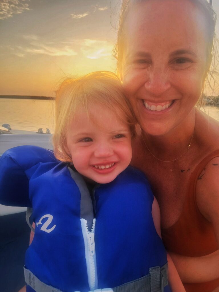 A smiling adult and child wearing a blue life jacket pose together on a boat at sunset, with warm sunlight and calm water in the background.