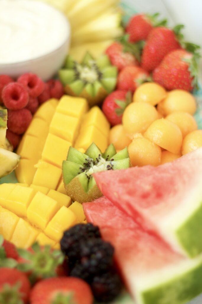 A colorful assortment of fresh fruits, including watermelon, strawberries, mango, raspberries, blackberries, cantaloupe, kiwi, and pineapple, arranged on a platter with a bowl of creamy dip in the background.