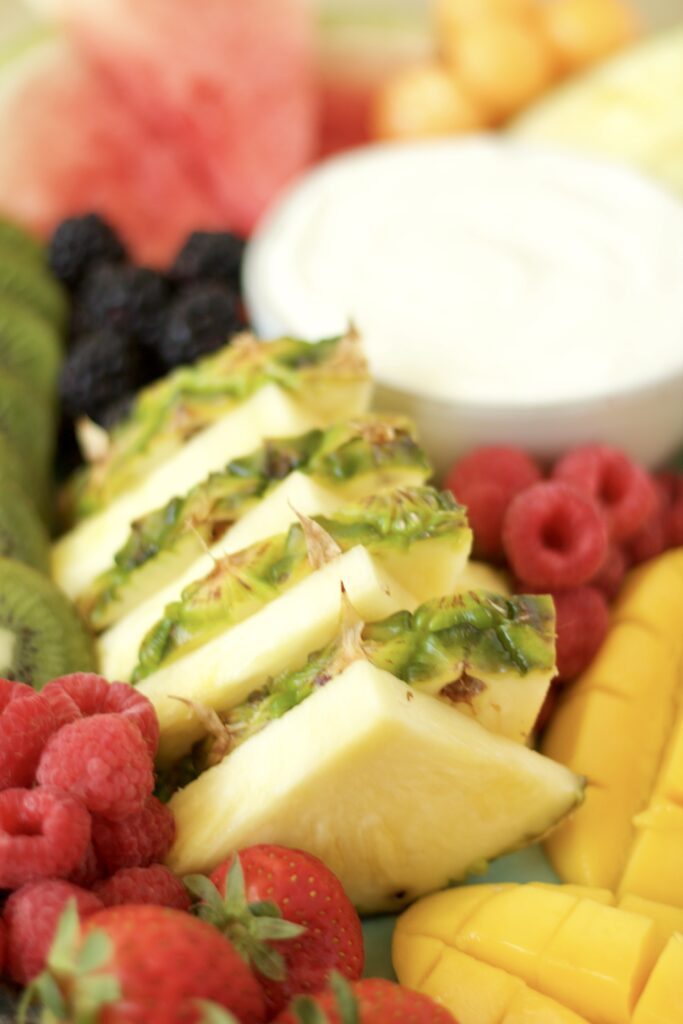 Sliced pineapple, raspberries, strawberries, blackberries, mango, kiwi, and cantaloupe are arranged around a bowl of white dip on a platter.