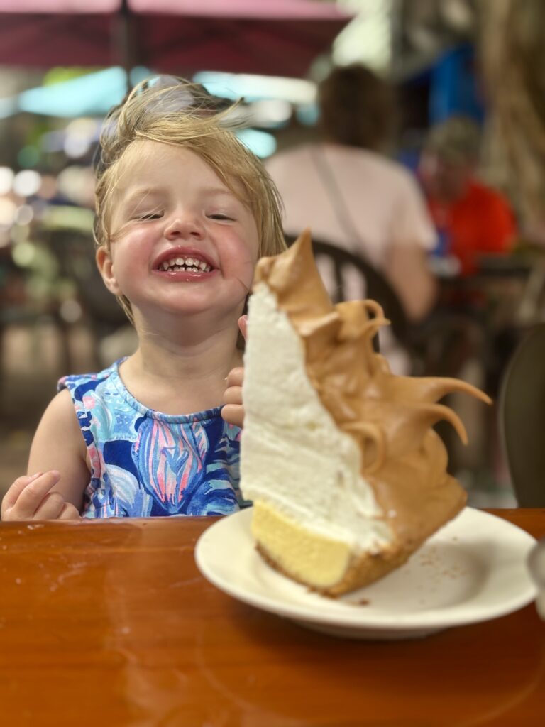 A smiling child in a blue dress sits at a table outdoors, excitedly looking at a large slice of pie with fluffy meringue topping on a plate in front of them.