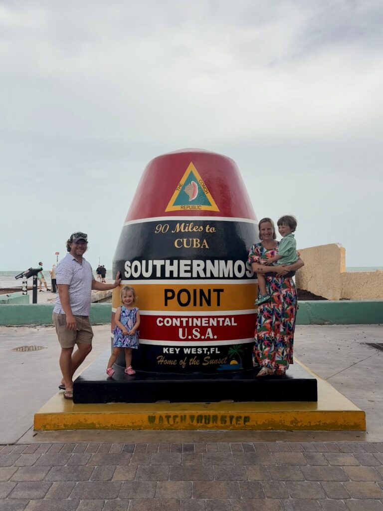 A family of four poses by the Southernmost Point buoy in Key West, Florida, with the ocean in the background. The buoy reads, 90 Miles to Cuba, Southernmost Point, Continental U.S.A..