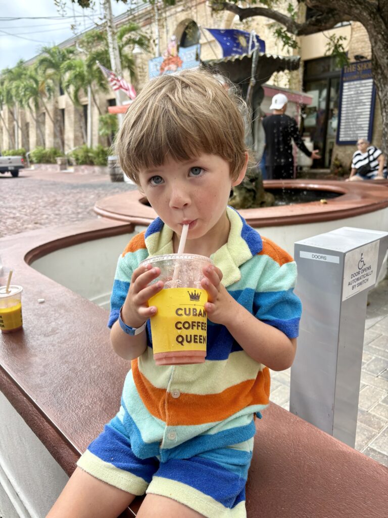 Young child in a brightly striped outfit sits on a bench, sipping a drink from a yellow Cuban Coffee Queen cup with a straw. Palm trees and street scenes are in the background.