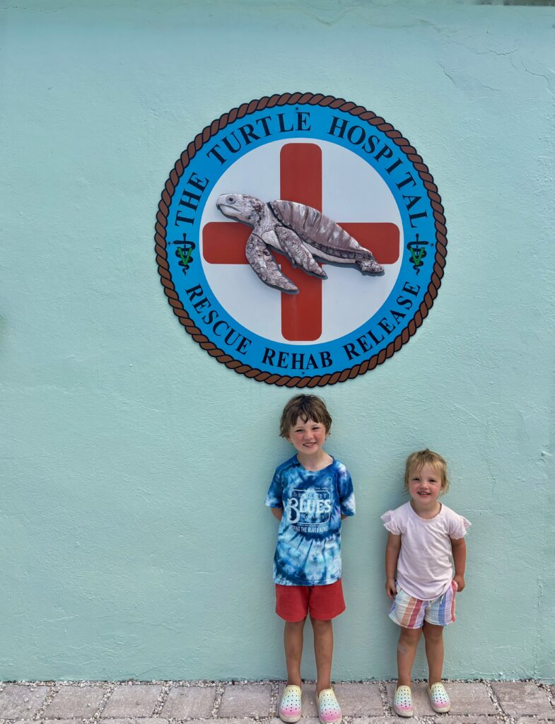 Two young children stand smiling in front of a mint green wall with a large round sign for The Turtle Hospital: Rescue Rehab Release featuring a turtle image and a red cross.