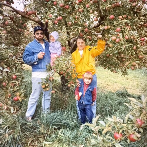 Four people and a child stand under an apple tree in a grassy orchard. The adults wear jackets, and two children, one wearing red, are picking apples. The tree is full of red apples and green leaves.