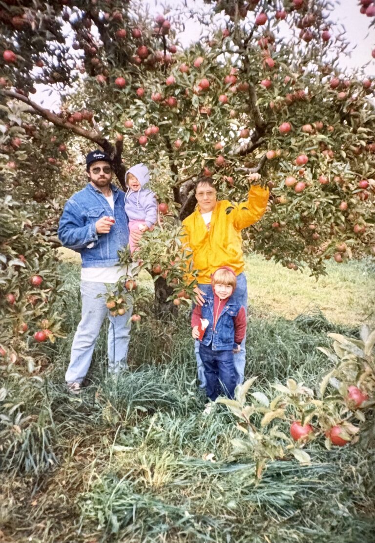 Four people and a child stand under an apple tree in a grassy orchard. The adults wear jackets, and two children, one wearing red, are picking apples. The tree is full of red apples and green leaves.