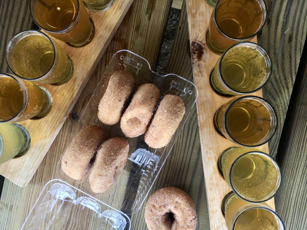 Six cinnamon sugar donuts in a plastic tray are surrounded by two wooden trays, each holding four glasses of amber-colored cider, on a wooden table.