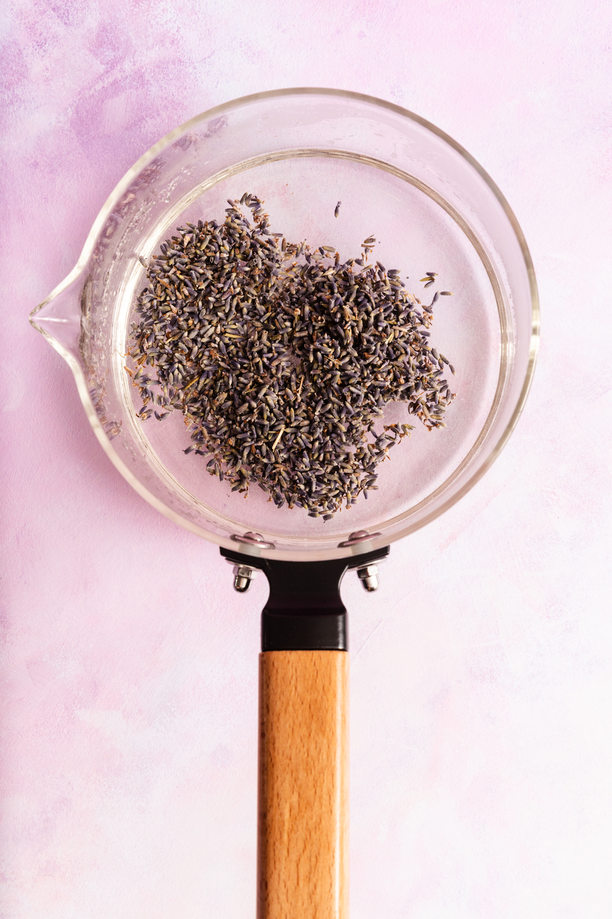 A clear glass saucepan with dried lavender flowers inside, seen from above, against a pastel pink background.
