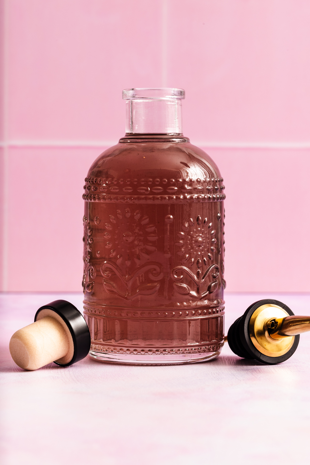 A decorative glass bottle filled with a brown liquid sits on a pink surface, with its cork and gold-tipped stopper placed beside it. The background features pink tiles.