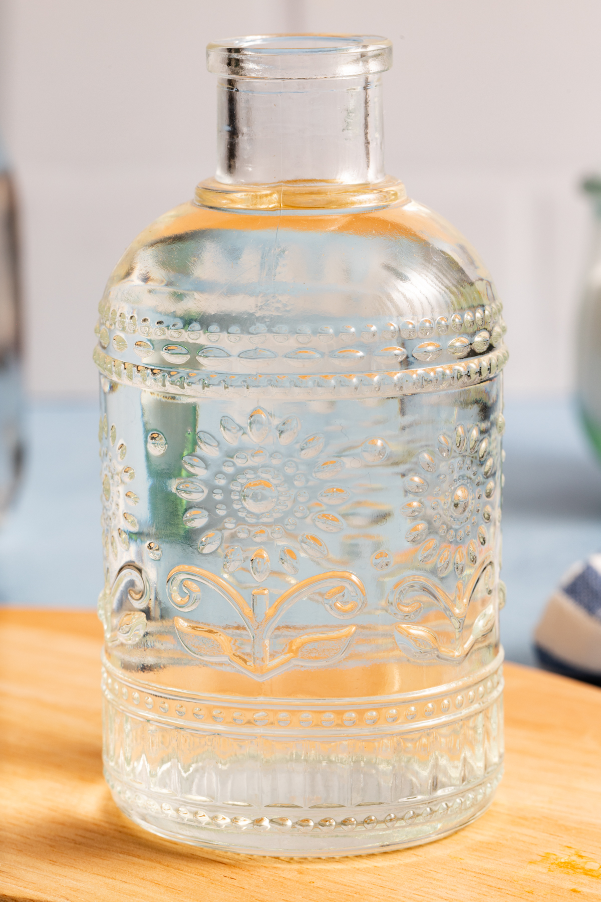 Clear glass bottle with embossed floral and geometric patterns, filled with a transparent liquid—perhaps the perfect result from learning how to make simple syrup—placed on a wooden surface with a soft-focus background.