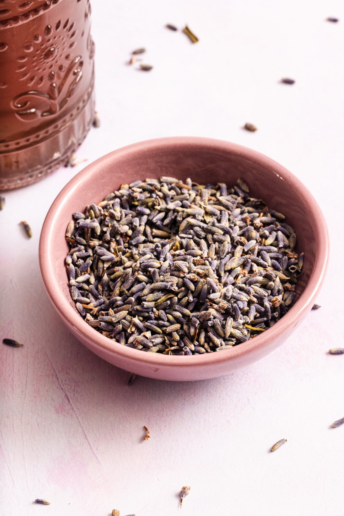 A pink bowl filled with dried lavender buds sits on a light surface. Some lavender pieces are scattered around the bowl, and part of an embossed container is visible in the upper left corner.