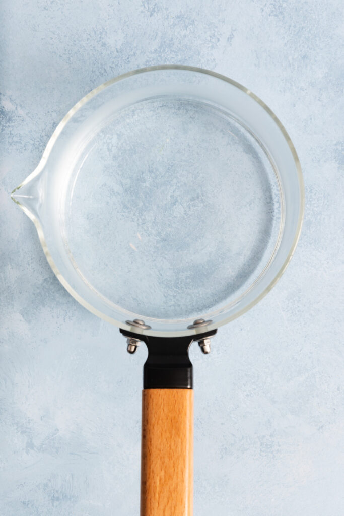 A clear glass saucepan with a wooden handle is placed on a light blue textured surface, viewed from above—an ideal setup if you’re learning how to make simple syrup at home.