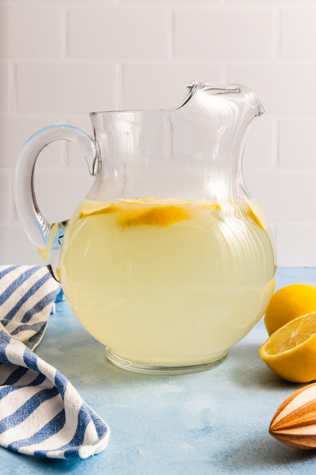 A glass pitcher of lemonade with lemon slices sits on a blue surface near a halved lemon, a striped towel, and a citrus juicer, against a white tiled background.