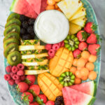 A platter with sliced watermelon, pineapple, mango, kiwi, strawberries, raspberries, blackberries, cantaloupe balls, and a bowl of white dip in the center, arranged on a green oval plate.